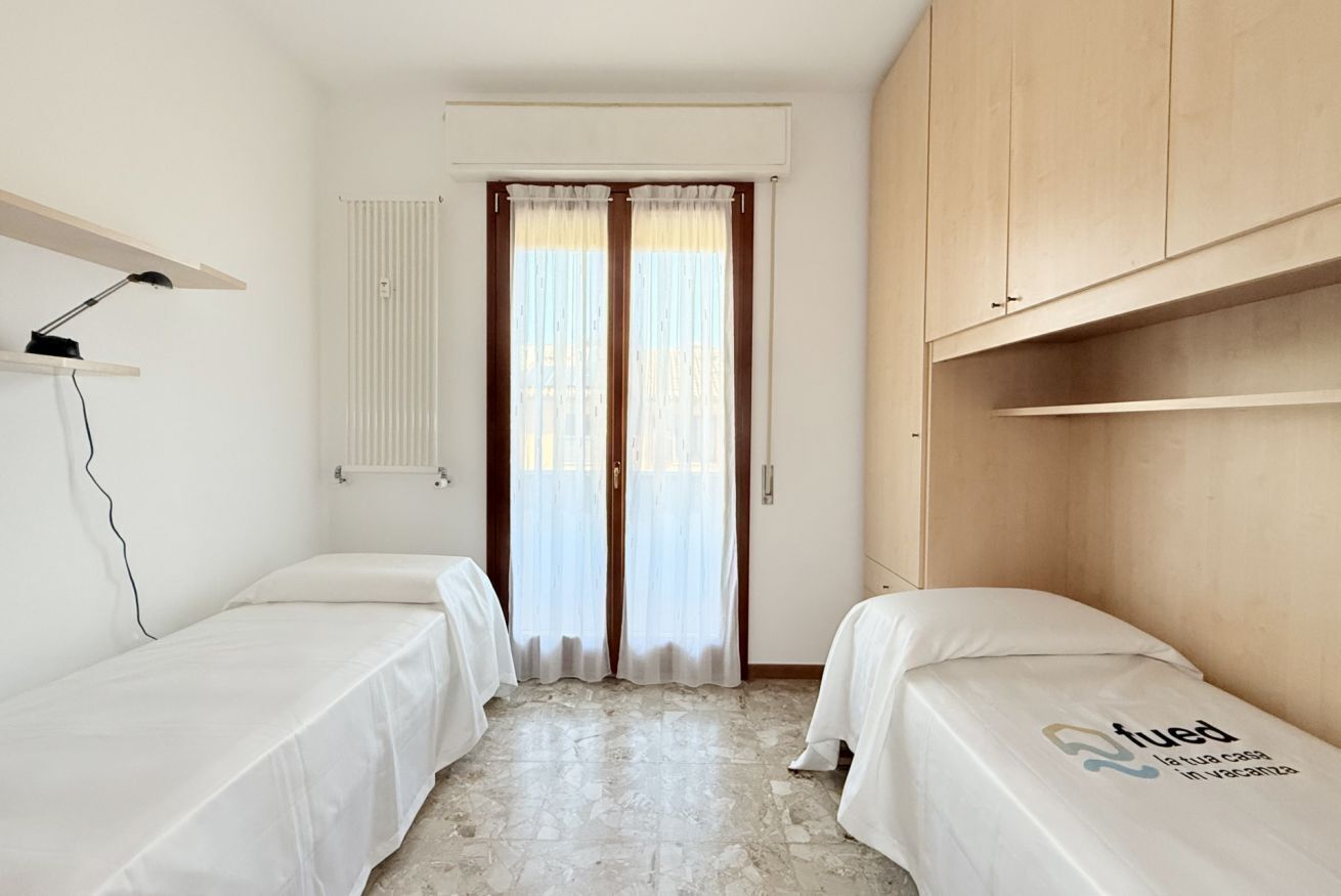 A small, clean room with two single beds, a desk with a lamp, a radiator, and a window with curtains.