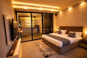 A modern hotel room with a large bed, a TV, a glass door leading to a bathroom, and a small desk with a lamp. - The Luxury Penthouse IGI Airport Delhi (New Delhi)