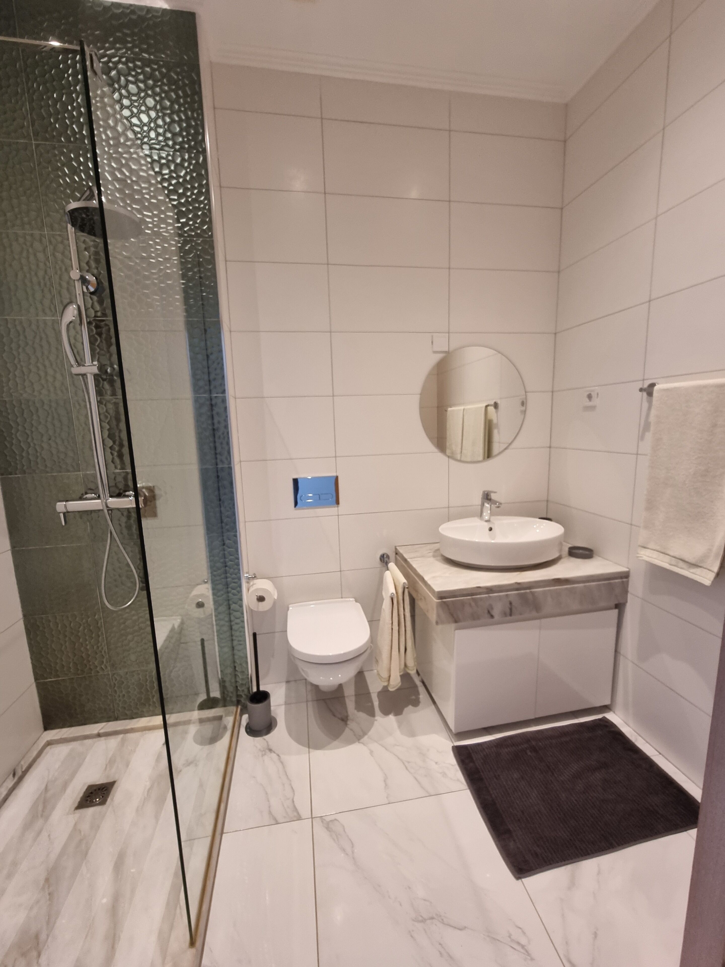 A modern bathroom with a glass shower enclosure, a white toilet, a sink with a mirror, and a dark floor mat.