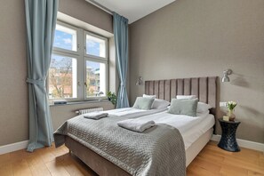 Une chambre à coucher avec un lit, une fenêtre avec des rideaux, une lampe de chevet et une petite table sur laquelle est posé un vase.