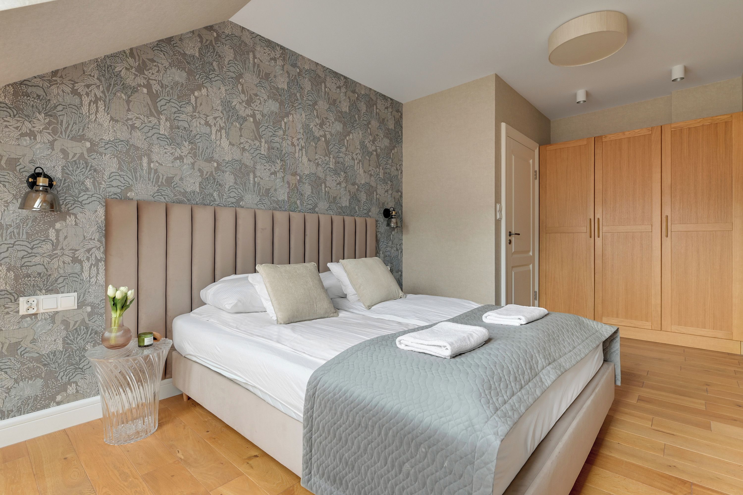 A bedroom with a bed, a nightstand, a wall-mounted lamp, a wardrobe, and a patterned wallpaper.