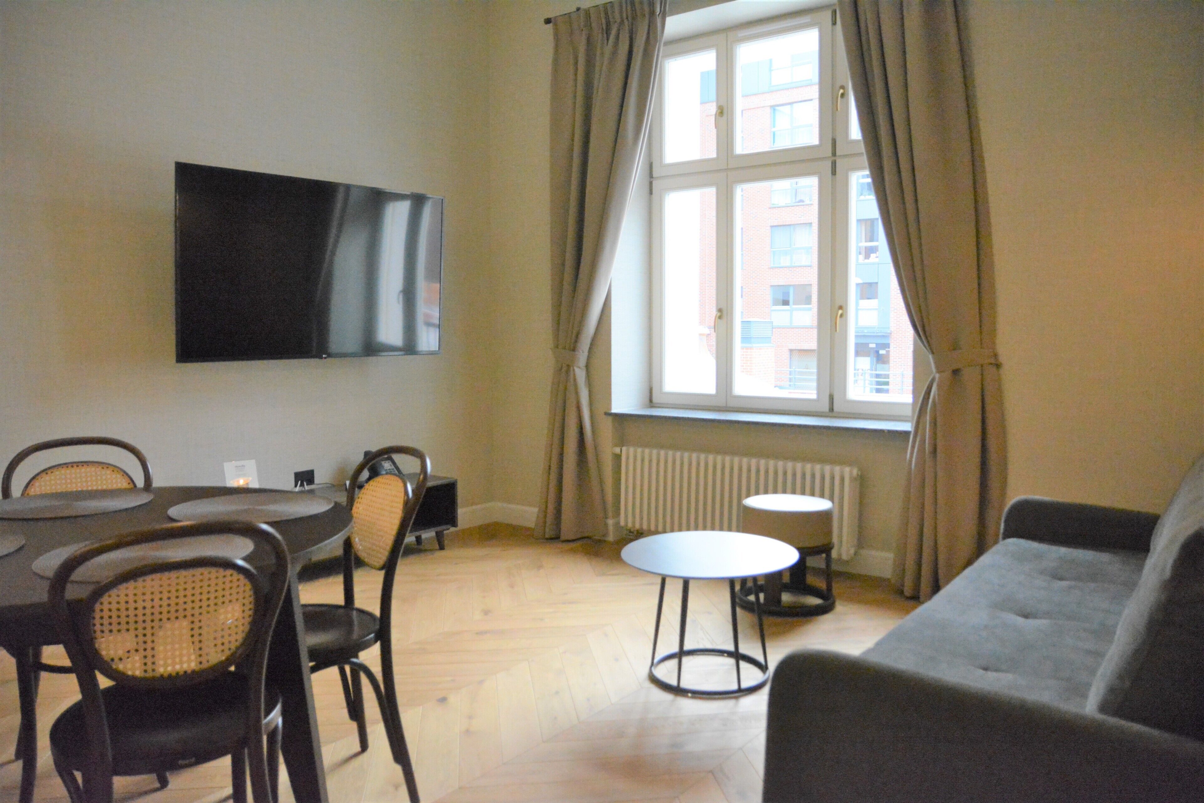 A room with a dining table, chairs, a sofa, a TV mounted on the wall, and a window with curtains.