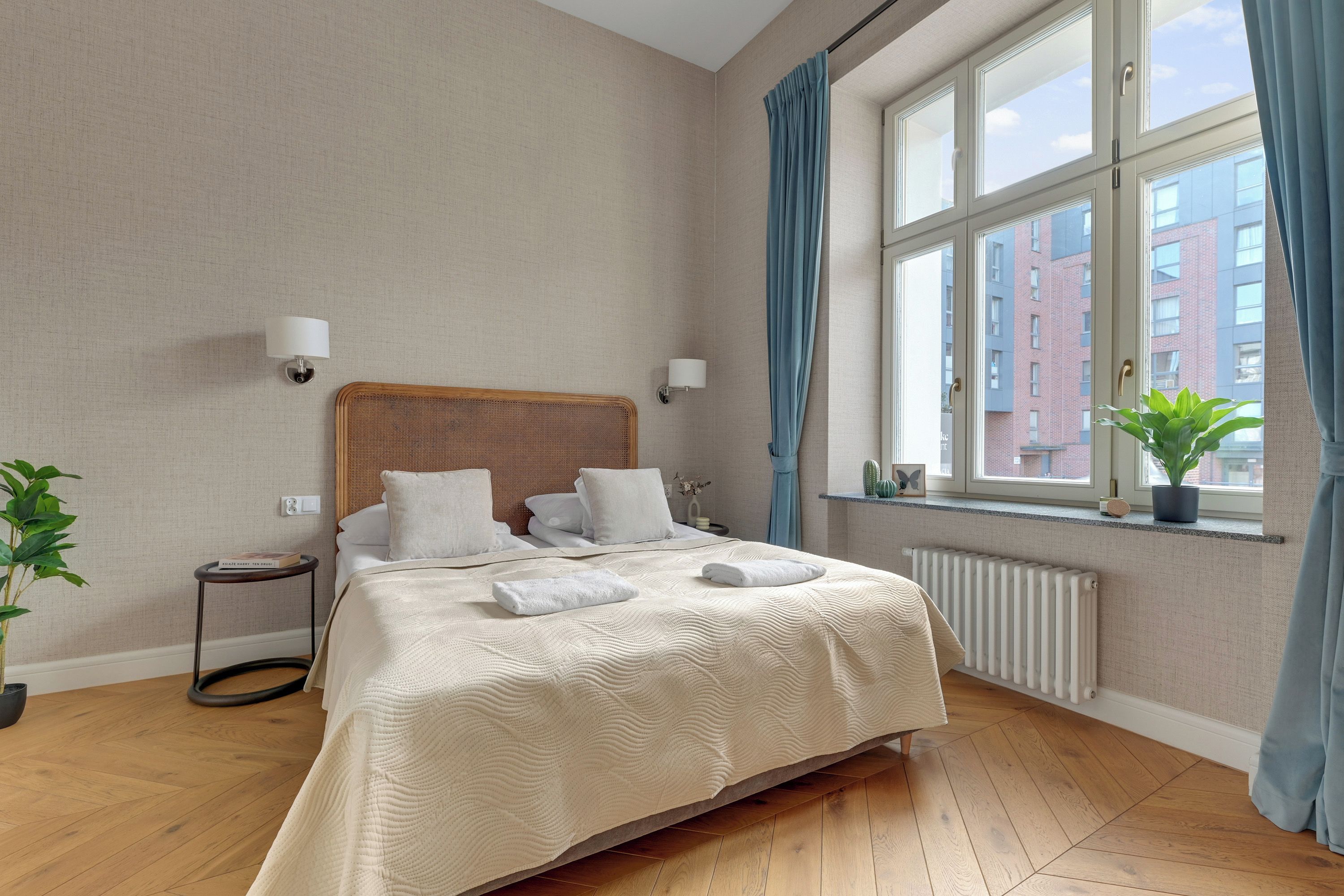 Ein Hotelzimmer mit einem großen Bett, zwei Nachttischlampen, einem Fenster mit Vorhängen und Blick auf umliegende Gebäude.