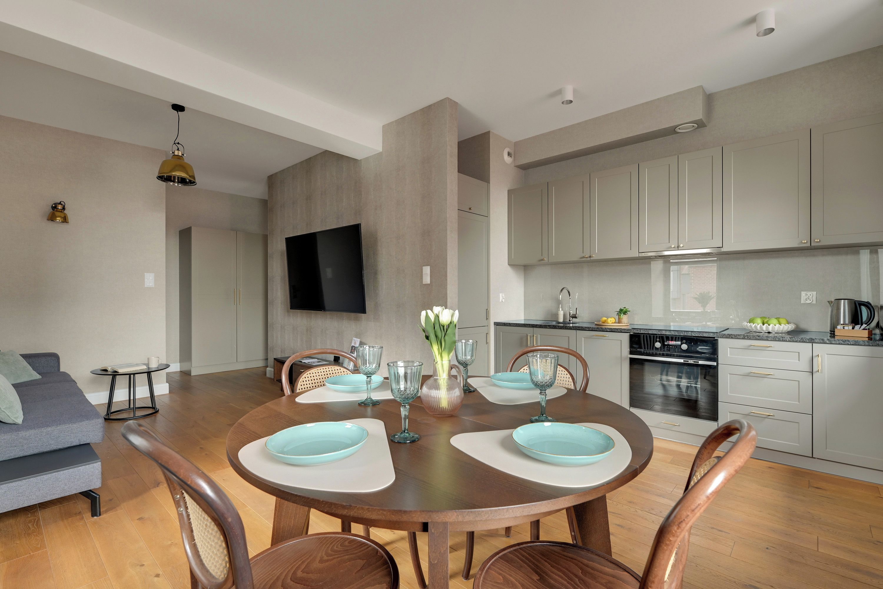 A modern kitchen with grey cabinets, a dining table set with blue plates, and a flat-screen TV mounted on the wall.