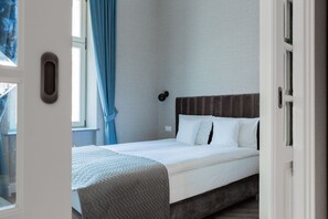 Una habitación de hotel con cama, ventana con cortinas, luz empotrada en la pared y una puerta.