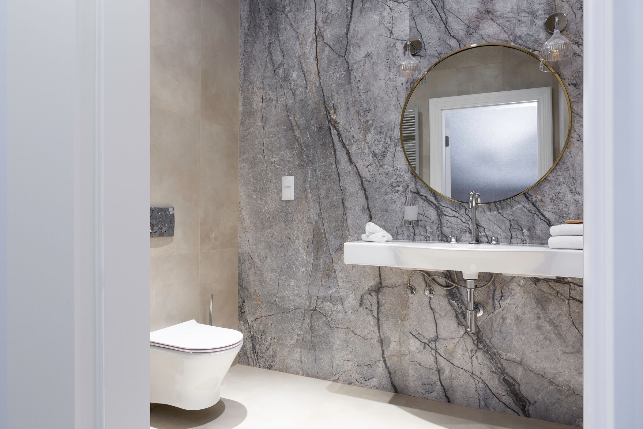 Un baño con pared de mármol, un espejo redondo, un lavamanos blanco y un inodoro.