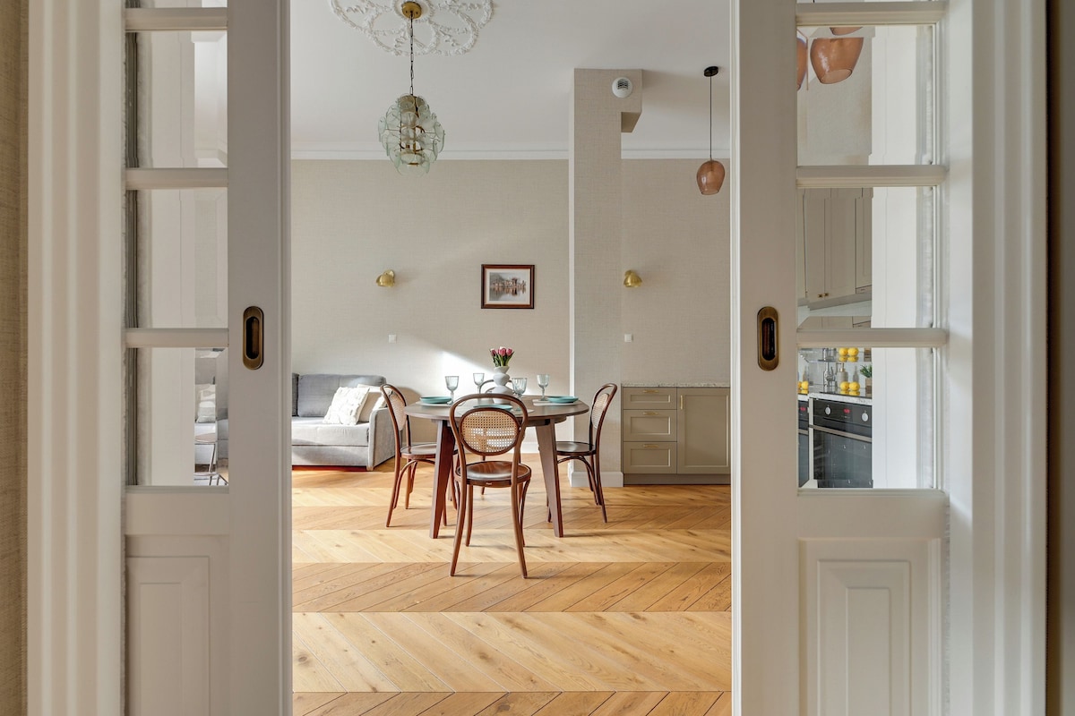 Une porte ouverte donne sur un espace repas doté d’un parquet en bois disposé en point de Hongrie, d’une table ronde et de deux chaises.