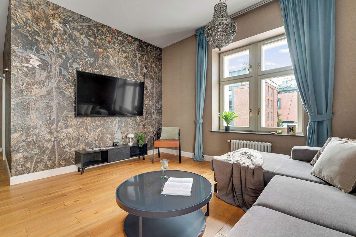Un salon moderne avec un téléviseur à écran plat, une table basse ronde sur laquelle repose un livre, un canapé et une fenêtre avec des rideaux.