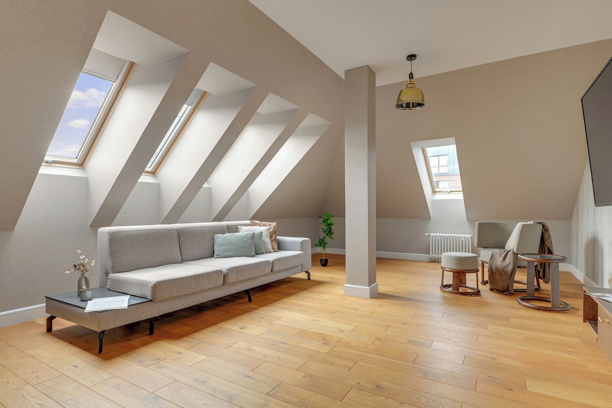 Un appartement moderne de style « loft », avec un plafond haut, des fenêtres de toit, un canapé gris, une petite table, une chaise et un radiateur.