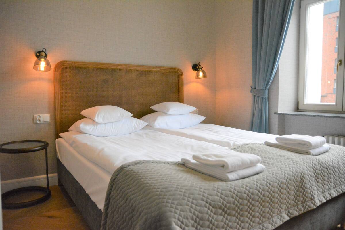 Quarto de hotel com cama de casal, duas mesas de cabeceira, janela com cortinas e luminárias fixadas na parede.