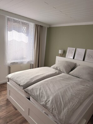 A bedroom with a bed, a window with curtains, a bedside table, and a wall with framed pictures. - Vacation apartment Börnecker Landruhe (Blankenburg (Harz))