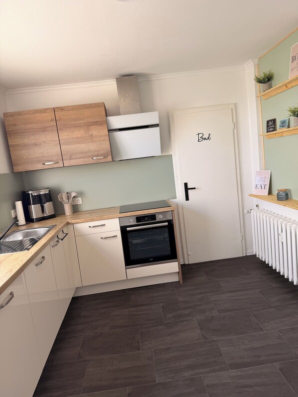 A compact kitchen with white cabinets, a built-in oven, a coffee maker, and a radiator. - Vacation apartment Börnecker Landruhe (Blankenburg (Harz))