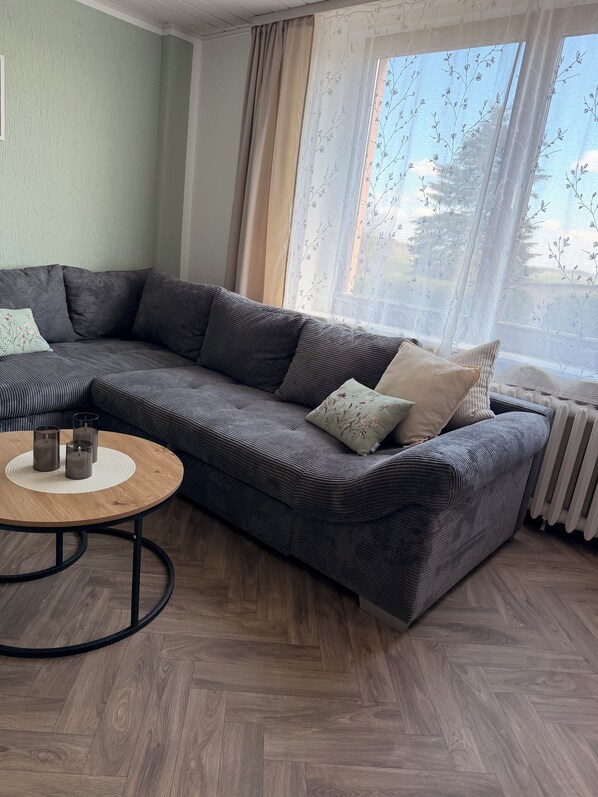 A modern living room with a sectional sofa, a round coffee table, and a large window with sheer curtains. - Vacation apartment Börnecker Landruhe (Blankenburg (Harz))