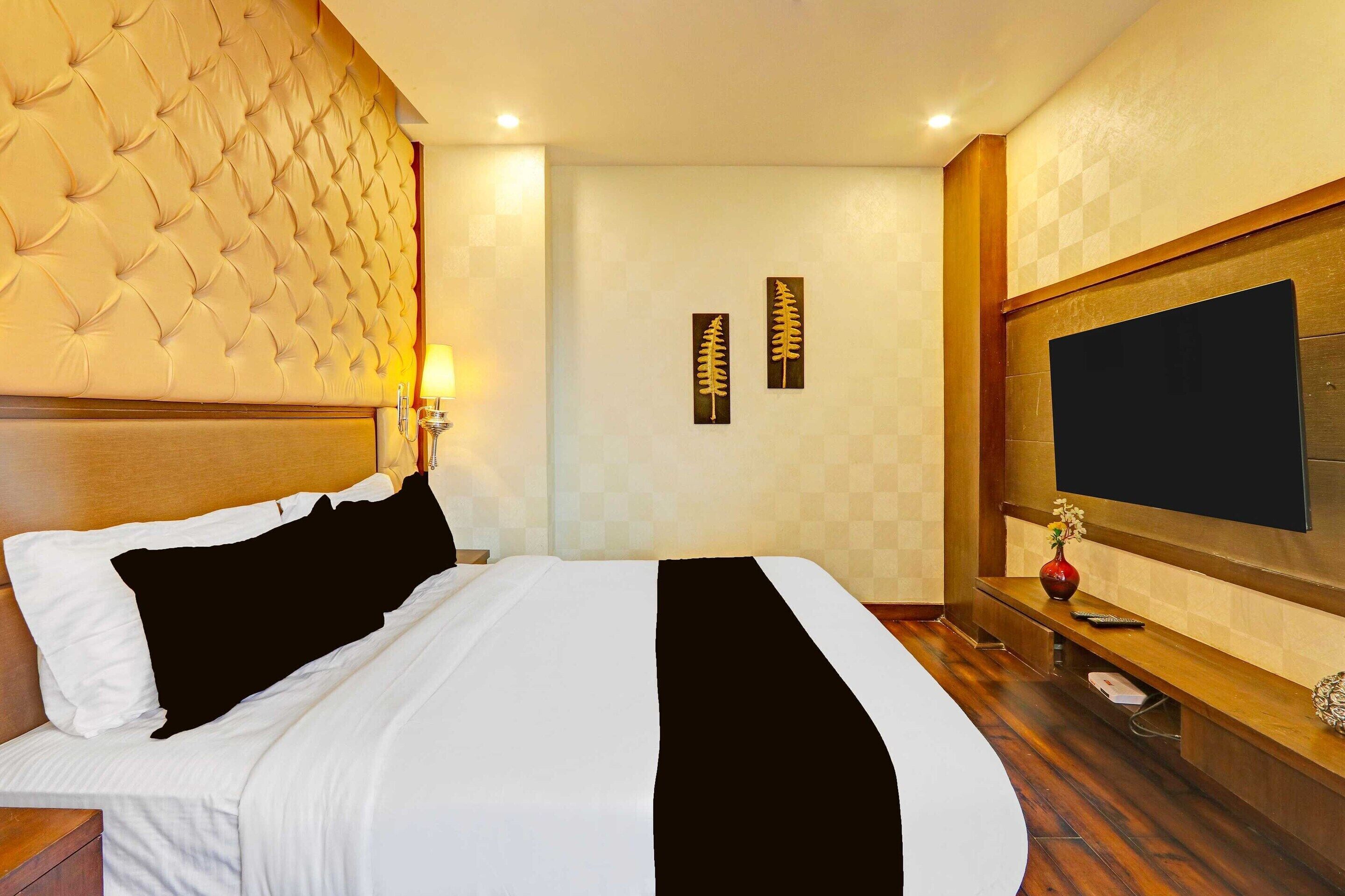 A hotel room with a tufted headboard, a flat-screen TV mounted on the wall, a bedside table with a vase, and a bed with white and black bedding.