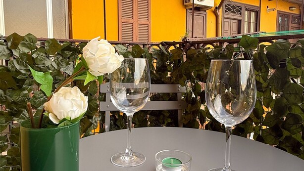 Une table avec deux verres à vin et un chandelier, entourée de verdure et avec un bâtiment jaune en arrière-plan.