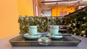 Une table avec deux tasses en céramique et un récipient en verre, placée devant un mur jaune orné d’une rambarde en métal et de feuillage vert.