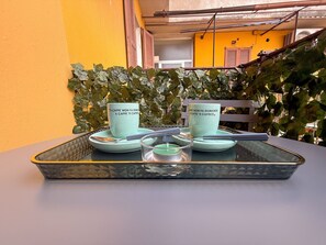 Ein Tisch mit zwei Keramiktassen und einem Glasbehälter, vor einer gelben Wand mit Metallgeländer und Grünpflanzen.