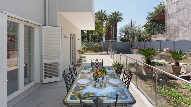 An outdoor dining area with a table set for four, surrounded by chairs, on a balcony with a view of palm trees and a building in the background. - Villa Cappuccini – Lower Floor Apartment with Shared Terrace, Wi-Fi & Air Conditioning (Sant'Agnello)