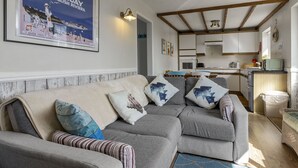A cozy living room with a grey sofa, a framed poster on the wall, and a kitchen in the background. - No.1 Cottage is a charming and cosy South Devon holiday cottage (Brixham)