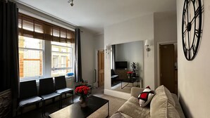 A living room with a sofa, coffee table, and chairs, a TV, and a large window with blinds. - Luxury Living at Charing Cross 26 | Best of London (London)