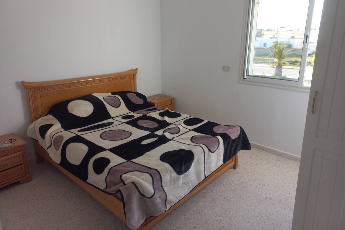 A bedroom with a wooden bed, a nightstand, a bedside table, and a window with a view of buildings and palm trees.