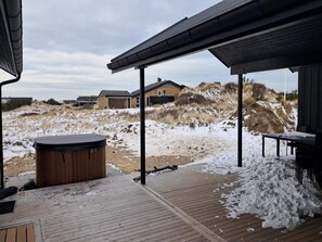 Eine Holzterrasse mit Whirlpool, Sitzgelegenheit im Freien und Blick auf Häuser und Dünen.