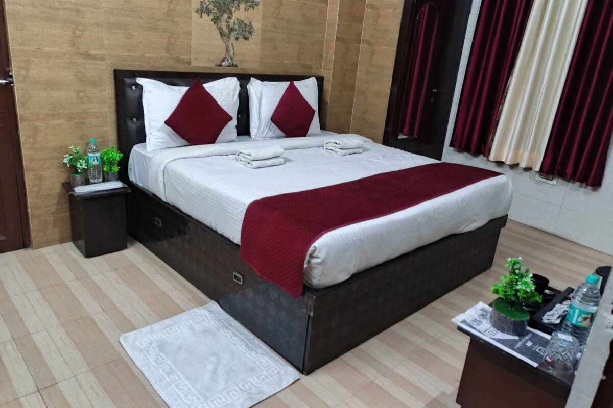 Quarto de hotel bem arrumado, com uma cama, mesa de cabeceira, uma pequena mesa com uma planta e uma janela com cortinas.