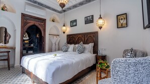 A bedroom with a large bed, a wooden headboard, a small side table, a chair, and a decorative wall with framed pictures. - Riad Nostalgia & Spa (Marrakech)