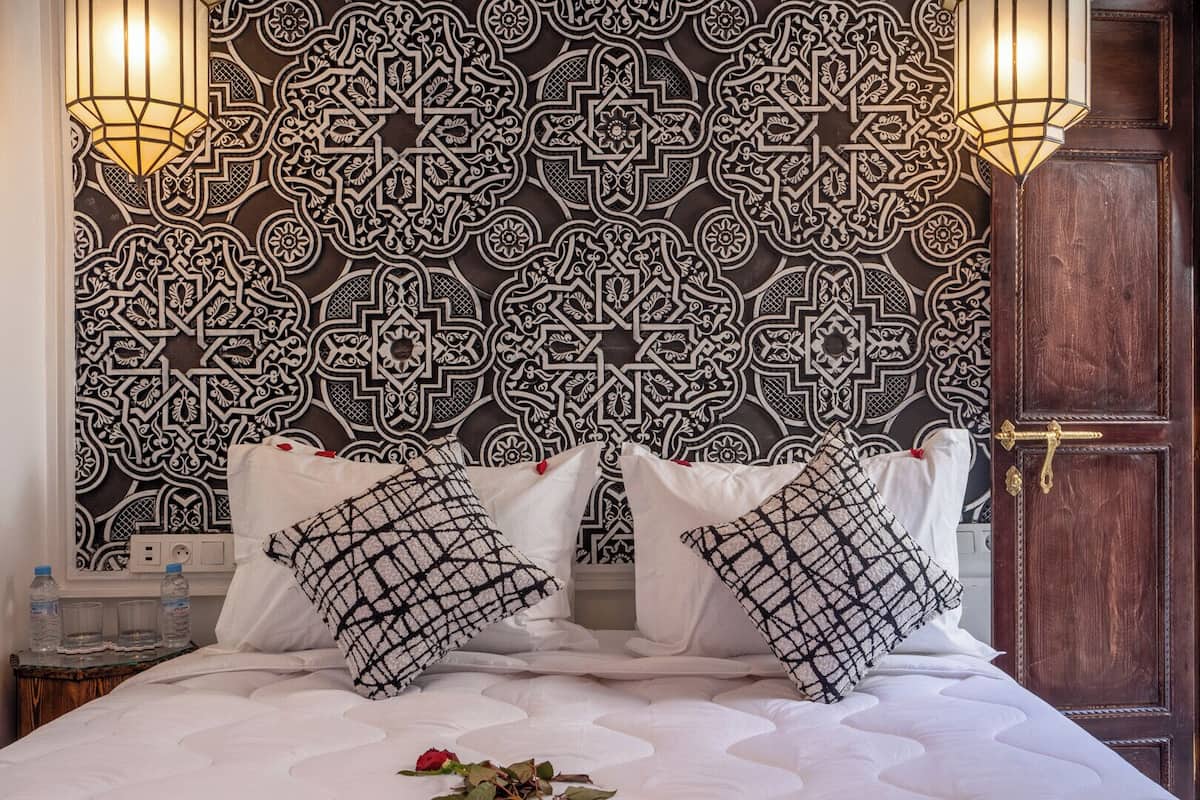 A bed with white bedding and black and white patterned pillows, a wooden door, and a wall with intricate black and white designs.