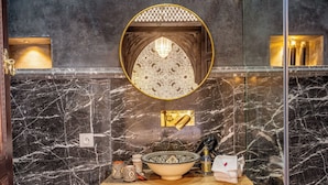 A bathroom with a marble wall, a round mirror, a sink with a black faucet, and a wooden vanity. - Riad Nostalgia & Spa (Marrakech)