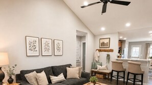 A living room with a dark grey sofa, a wooden coffee table, and a ceiling fan. There are framed botanical prints on the wall and a kitchen area in the background. - Peaceful 4-bedroom Fayetteville getaway near the University of Arkansas! Enjoy a heated & cooled game room, fenced backyard with playground, gas grill, and plenty of space for the whole family. (Fayetteville)