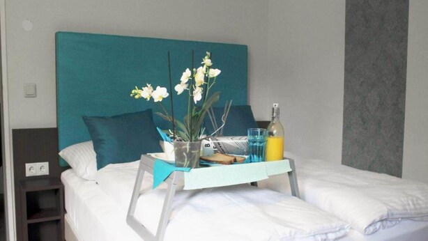 A neatly made bed with white linens, a teal headboard, a bedside table with a lamp, a tray with a bottle, a glass, and a potted orchid. - 2 Person Holiday Park Home in Bendorf (Bendorf)