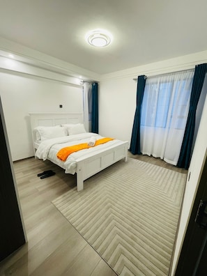 Ein Schlafzimmer mit Bett, Deckenleuchte, Fenster mit Vorhängen und einem Teppich mit Fischgrätmuster.