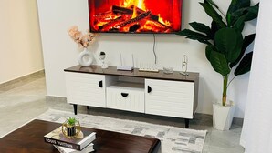 Uma sala de estar com uma TV exibindo uma lareira, um armário branco com gavetas, uma mesa de centro com um livro e um vaso de plantas.