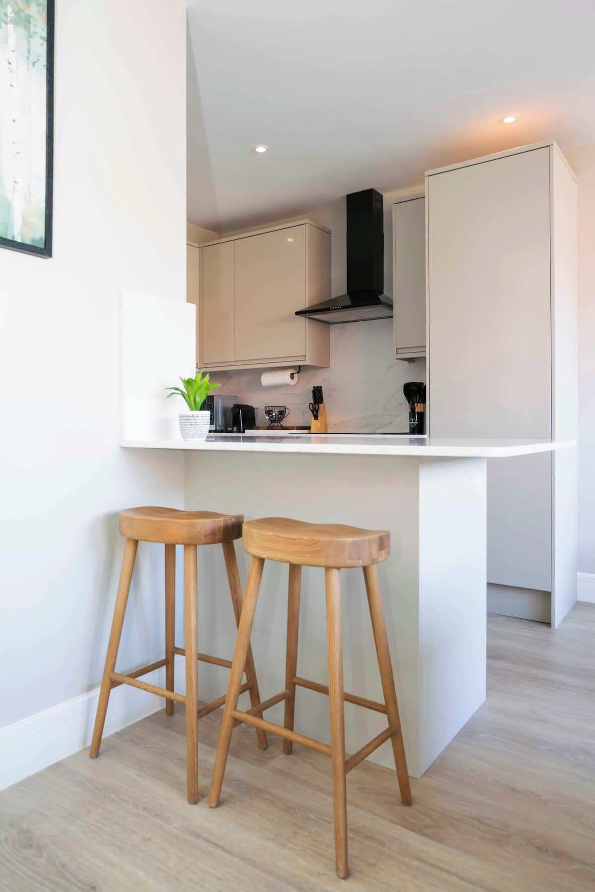 A modern kitchen with white cabinetry, a black range hood, and two wooden stools.