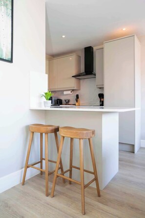 A modern kitchen with white cabinetry, a black range hood, and two wooden stools. - Wimbledon 2BR | Indoor-Outdoor Living & Patio (London)
