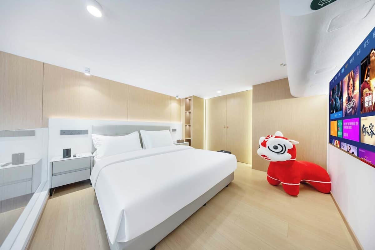 A modern hotel room with a large bed, bedside tables, a nightstand, a red plush toy, and a wall with a digital screen displaying colorful content.