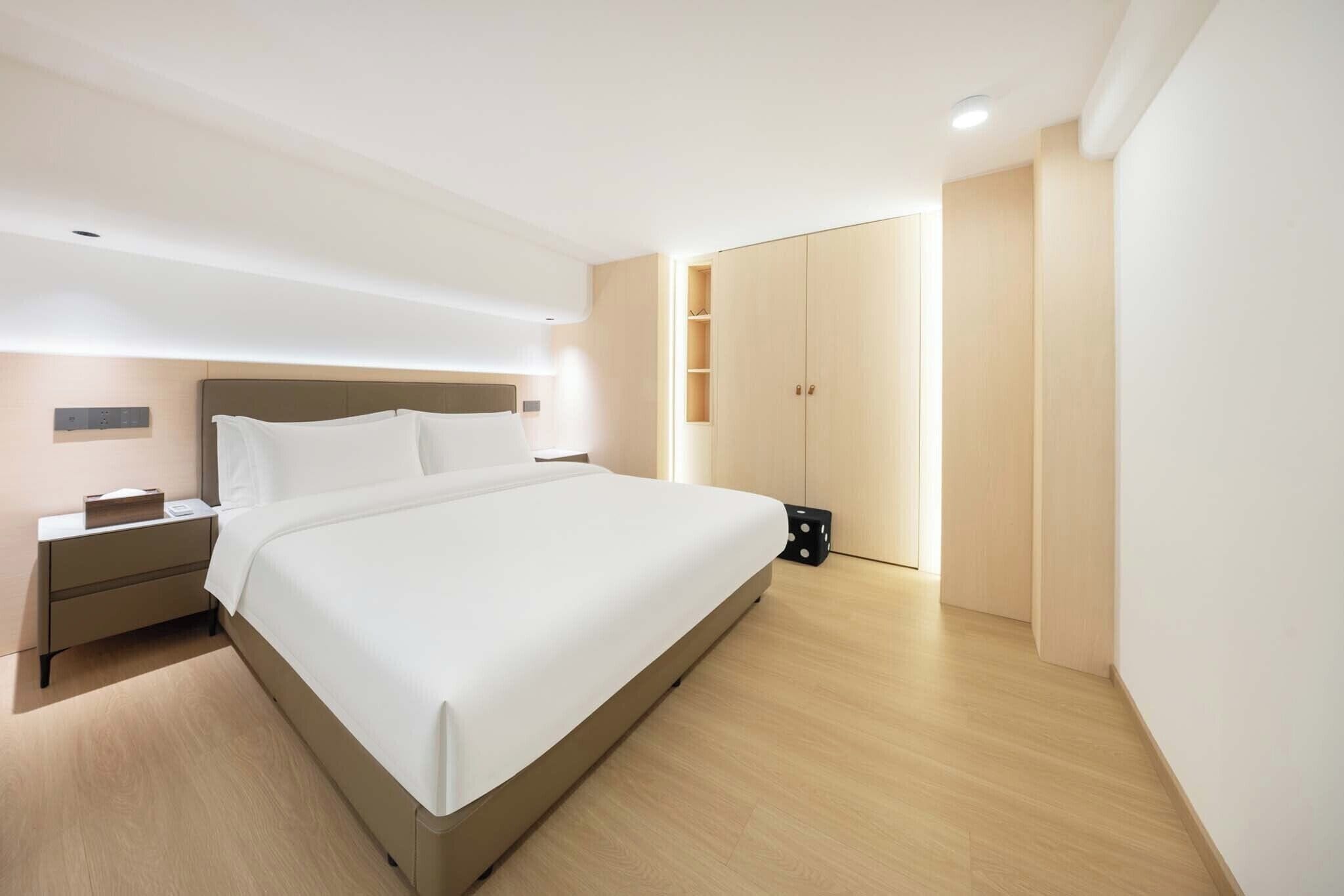 A modern hotel room with a large bed, bedside tables, a wardrobe, and a small seating area.