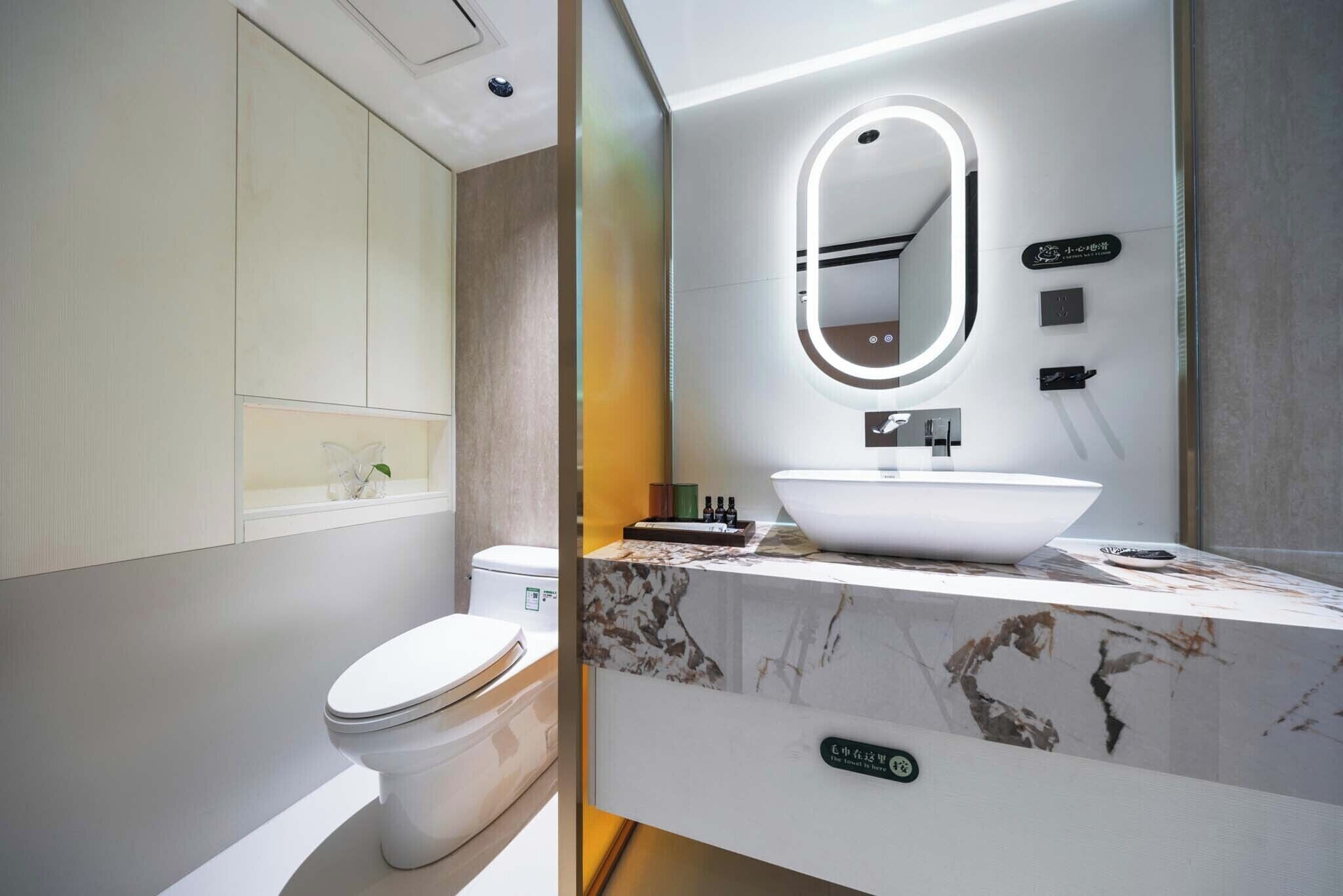 Un baño moderno con inodoro blanco, un espejo ovalado grande, lavamanos blanco y encimera de mármol.