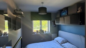 Un dormitorio con cama, ventana, radiador, repisa con objetos y televisor montado en la pared.