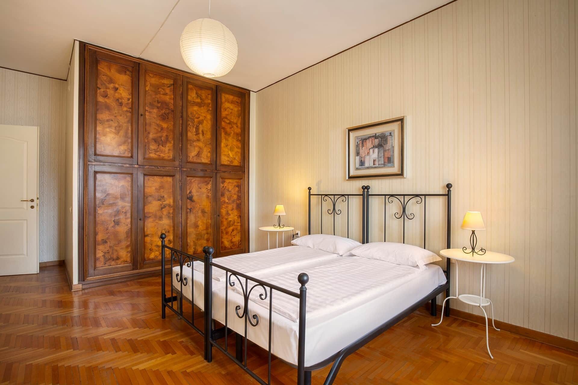 A bedroom with a wooden wardrobe, a bed with a metal frame, two bedside tables with lamps, and a hanging light fixture.