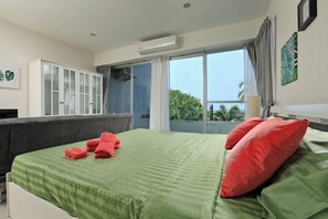 Ein Schlafzimmer mit Bett, grüner Bettwäsche, roten Kissen, einem grauen Sofa, einem weißen Kleiderschrank und einem großen Fenster mit Blick.