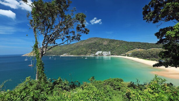 Paisagem costeira com uma praia, águas azuis cristalinas e uma colina com edifícios.