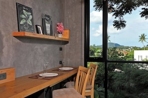 Ein Essbereich mit einem Holztisch, Stühlen und Blick auf Grünflächen und Gebäude durch große Fenster.