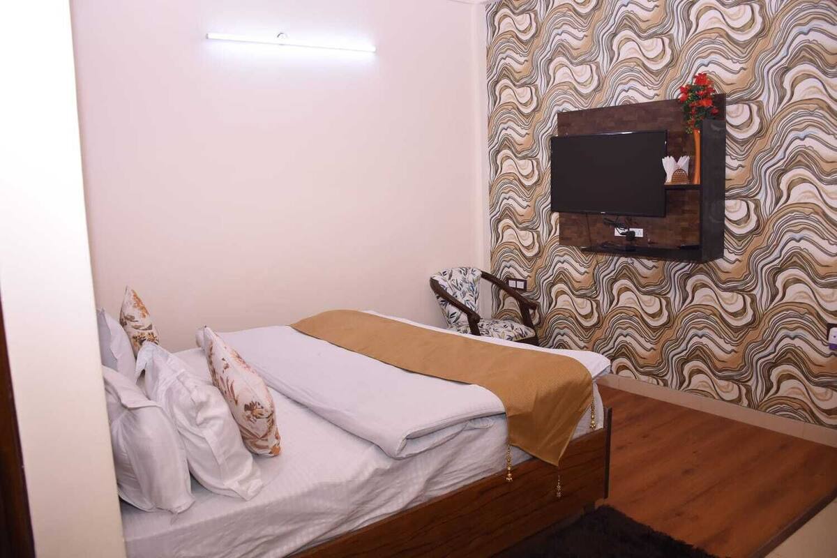 Una cama individual con una manta marrón, suelo de madera, pared estampada y un televisor montado.