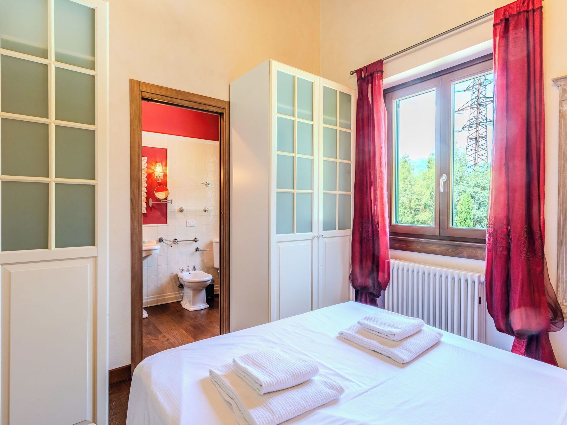 A bedroom with a bed, a window with curtains, a radiator, and a view of the outdoors.