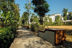 A pathway lined with greenery and a wooden bench, leading to a modern building with a flat roof and large windows. - Laguna Park | Bangtao Beach | 3-bedroom Townhouse (Bangtao)