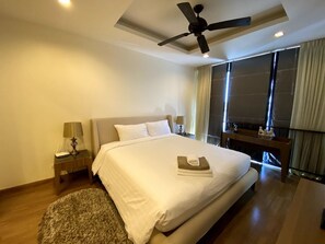 Ein Hotelzimmer mit einem Bett, einem Deckenventilator, einem Nachttisch mit Lampe, einem kleinen Tisch und Blick durch ein Fenster nach draußen.