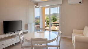 Una habitación con televisor, una mesa de comedor redonda, sillas blancas, un sofá blanco y un balcón con vistas al campo.