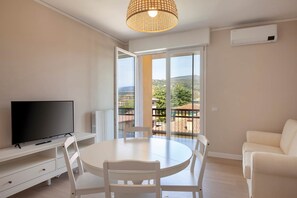 Ein Zimmer mit Fernseher, einem runden Esstisch, weißen Stühlen, einem weißen Sofa und einem Balkon mit Blick auf die Landschaft.